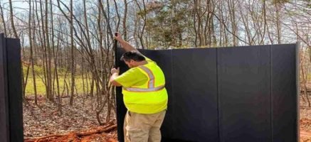 Worker installing a tall privacy fence panel outdoors wearing safety gear